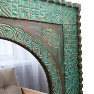 May include: A decorative mirror with a carved wooden frame. The frame features intricate patterns and is painted in shades of teal and reddish-brown. The mirror has an arched top and a detailed border.