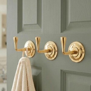 May include: Three brass towel hooks mounted on a sage green door. Each hook has a circular base with a decorative rope-like edge. A beige towel hangs from one of the hooks, adding a touch of warmth to the scene.