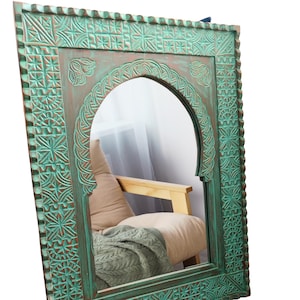 May include: A large, ornate, arched mirror with a distressed teal finish. The wooden frame features intricate carved geometric and floral patterns. The mirror reflects a glimpse of a chair with a pillow and a blanket.