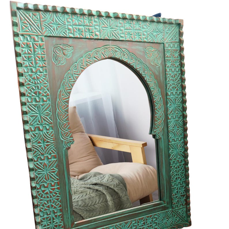 May include: A large, ornate, arched mirror with a distressed teal finish. The wooden frame features intricate carved geometric and floral patterns. The mirror reflects a glimpse of a chair with a pillow and a blanket.