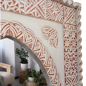 May include: A decorative, carved wooden mirror with a white frame and intricate, geometric patterns in a natural wood color. The mirror has an arched top and a scalloped edge.