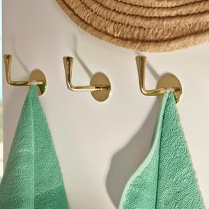 May include: Three gold-colored wall hooks with a curved design are mounted on a white wall. Two green towels hang from the hooks. A woven straw hat is partially visible at the top of the image.