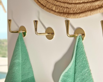 Könnte beinhalten: Drei goldfarbene Wandhaken mit geschwungenem Design sind an einer weißen Wand befestigt. Zwei grüne Handtücher hängen an den Haken. Ein gewebter Strohhut ist oben im Bild teilweise zu sehen.
