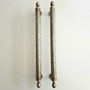 May include: Two antique brass door pulls with ornate, swirling patterns. Each pull is a long, cylindrical shape with a decorative finial at the top and bottom. The pulls are mounted on small, round plates.