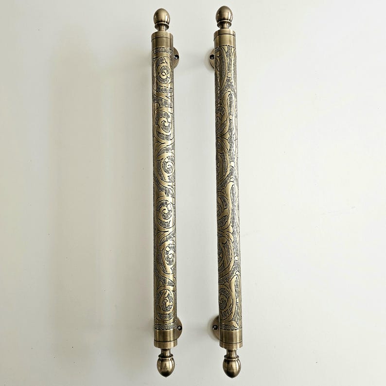 May include: Two antique brass door pulls with ornate, swirling patterns. Each pull is a long, cylindrical shape with a decorative finial at the top and bottom. The pulls are mounted on small, round plates.
