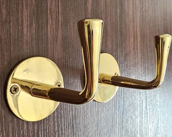 May include: Two brass-colored coat hooks with a dark wood accent. The hooks are mounted on round, gold-colored plates and attached to a wooden door. The hooks have a curved design.