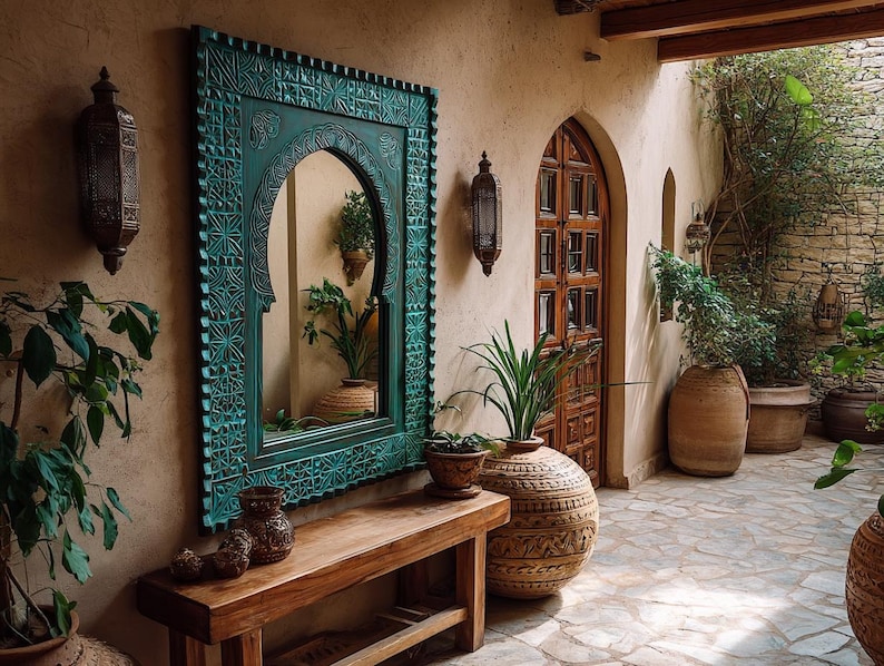 May include: An outdoor scene featuring a teal-framed mirror with intricate carvings, reflecting plants and a warm-toned interior. A wooden bench holds decorative vases. The space includes a wooden door, stone flooring, and potted plants, creating a serene, earthy ambiance.