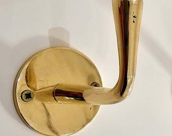 May include: A gold-colored wall hook with a rounded base and a curved hook. The hook is attached to the wall with two screws. The hook is made of metal and has a shiny finish.