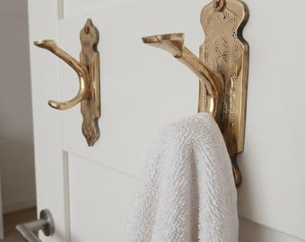 May include: Two ornate brass wall hooks with a white towel hanging on one. The hooks are mounted on a white door. The hooks have a decorative design and a curved arm for hanging items. The towel is white.