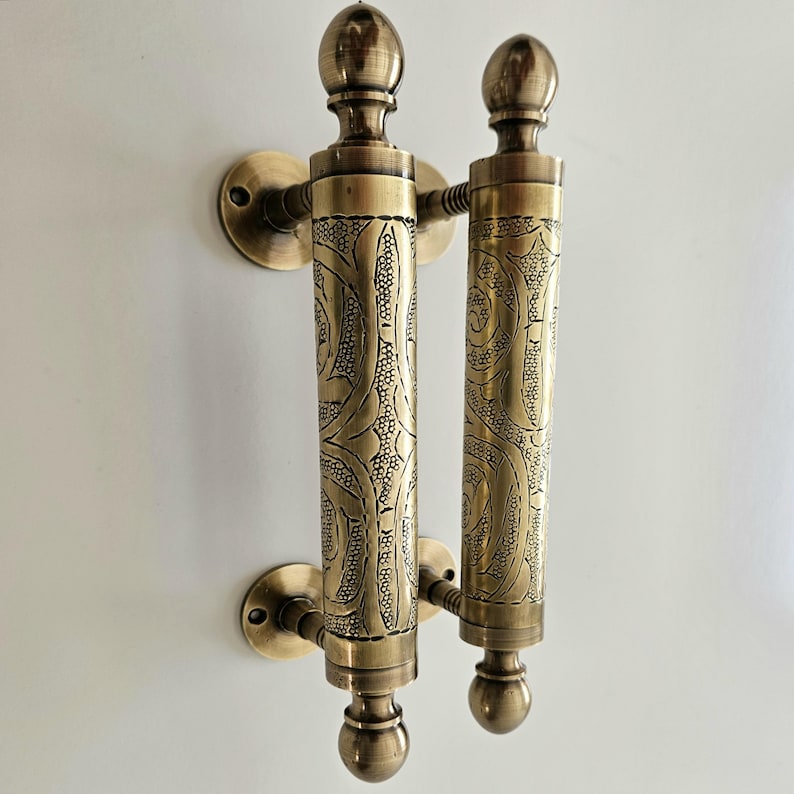 May include: Two antique brass door handles. Each handle has a cylindrical body with an ornate, textured pattern. The handles are mounted on round plates and topped with a decorative finial. The overall color is a warm, aged brass.