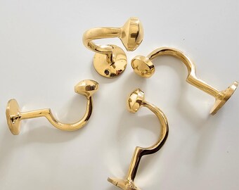 May include: Four brass curtain tieback hooks. Each hook has a curved arm and a rounded end. The hooks are a shiny gold color and are designed to be mounted on a wall. The hooks are made of metal and are likely vintage or antique.