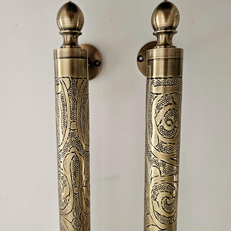May include: Two antique brass door handles with ornate floral and swirl patterns. Each handle has a cylindrical body with detailed engravings and a rounded top. The handles are mounted on circular backplates.