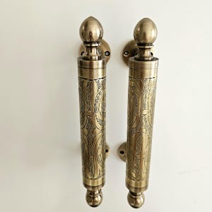 May include: Two antique brass door handles with intricate floral patterns. Each handle features a cylindrical grip with detailed engravings and a decorative finial at the top and bottom. The handles are mounted on round backplates.