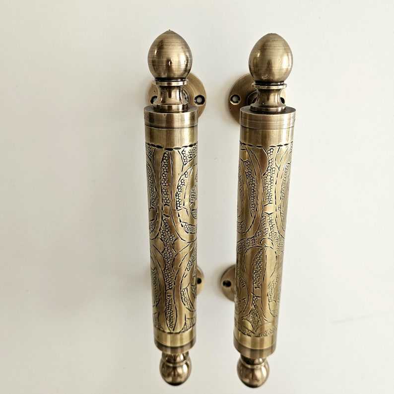 May include: Two antique brass door handles with intricate floral patterns. Each handle features a cylindrical grip with detailed engravings and a decorative finial at the top and bottom. The handles are mounted on round backplates.