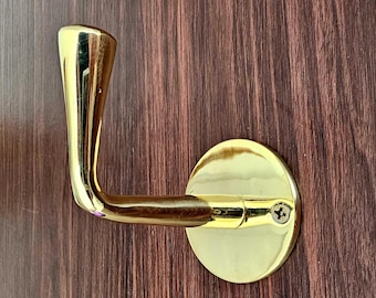 May include: A gold-colored door handle with a curved design. The handle is attached to a round, gold-colored plate with a single screw. The background is a dark brown wooden door.