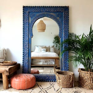 May include: A large, blue, ornate mirror with an arched top and carved details. The mirror reflects a bedroom scene with a bed, bench, and woven basket. The mirror is framed by a detailed border, adding a touch of bohemian style to the room.