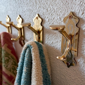 May include: Close-up of four ornate brass wall hooks. The hooks are mounted on a textured white wall, with two colorful towels hanging from them. The hooks have a decorative design and a curved shape.