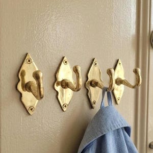 Puede incluir: Cuatro perchas de pared de latón con placas traseras en forma de diamante están montadas en una puerta beige. Una prenda azul cuelga de uno de los ganchos. Estas perchas son un diseño clásico para colgar abrigos, sombreros u otros objetos.