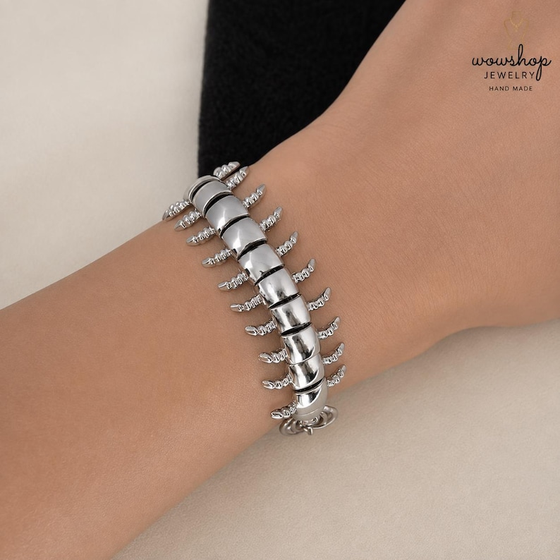 May include: A silver-toned bracelet designed to resemble a centipede, featuring a segmented body with multiple small, curved legs extending from each side. The metallic links are polished and smooth, creating a continuous, articulated structure.