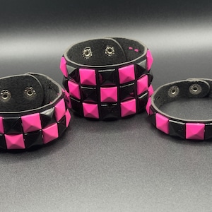 Op de afbeelding: Drie zwarte leren armbanden met afwisselende rijen piramidevormige studs in felroze en zwart. Elke armband is voorzien van zilverkleurige metalen drukknopen aan de binnenzijde, met variërende breedtes van één tot drie rijen met studs.