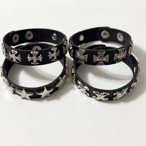 May include: Four black leather bracelets with silver-colored embellishments. Two bracelets feature cross designs, one has star studs, and the other has an X pattern. Each bracelet has a snap closure.