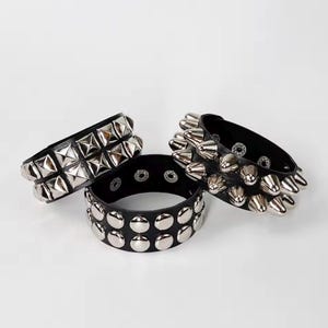Puede incluir: Tres pulseras de cuero negro con tachuelas de metal plateado. Una tiene tachuelas en forma de pirámide, otra tachuelas redondeadas y la tercera tachuelas cónicas. Las pulseras se muestran sobre una superficie blanca.