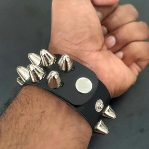 Puede incluir: Brazalete de cuero negro con p&uacute;as plateadas en forma de cono. La pulsera tiene un cierre de bot&oacute;n a presi&oacute;n y un detalle de bot&oacute;n plateado. Las p&uacute;as est&aacute;n espaciadas uniformemente.