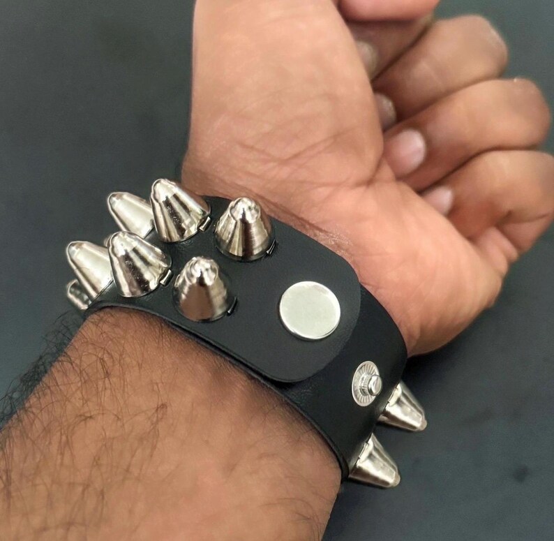 Puede incluir: Brazalete de cuero negro con p&uacute;as plateadas en forma de cono. La pulsera tiene un cierre de bot&oacute;n a presi&oacute;n y un detalle de bot&oacute;n plateado. Las p&uacute;as est&aacute;n espaciadas uniformemente.