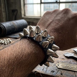 May include: Black leather bracelet with silver cone studs. The bracelet is a bold accessory, featuring rows of metallic spikes. The design is edgy and suitable for alternative fashion styles. The bracelet is a statement piece.