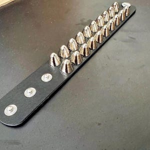 Puede incluir: Una pulsera de cuero negro con dos filas de tachuelas c&oacute;nicas plateadas. La pulsera tiene cierres de bot&oacute;n a presi&oacute;n plateados. Las tachuelas est&aacute;n espaciadas uniformemente y tienen un acabado met&aacute;lico brillante. La pulsera se muestra sobre una superficie oscura.