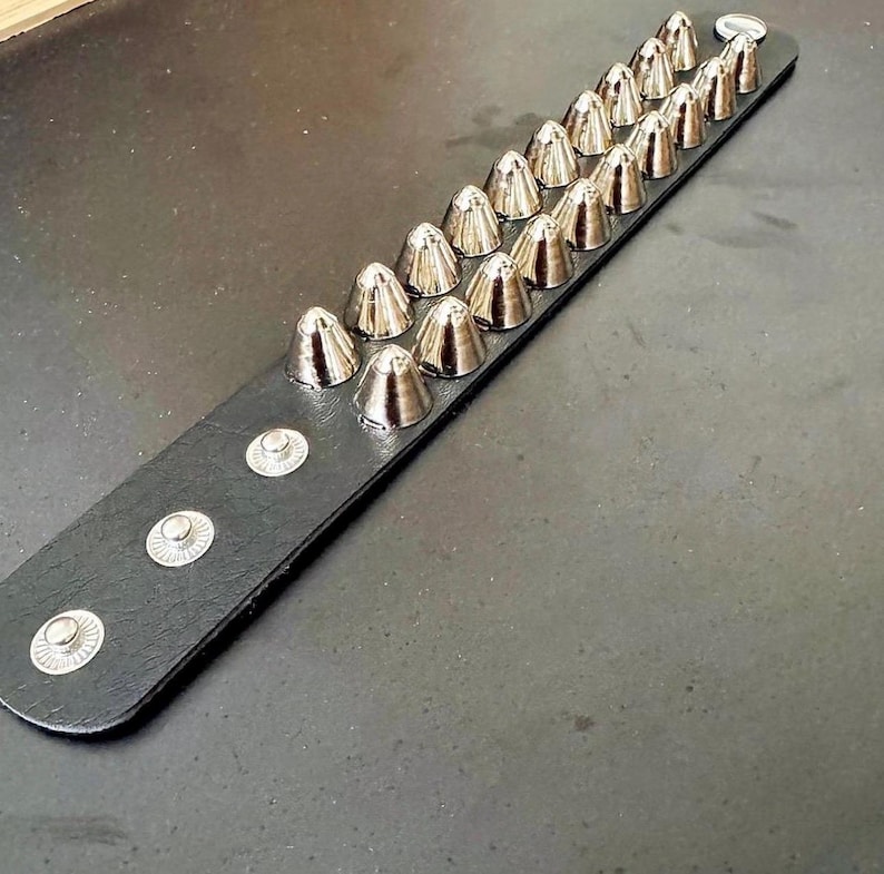 Puede incluir: Una pulsera de cuero negro con dos filas de tachuelas c&oacute;nicas plateadas. La pulsera tiene cierres de bot&oacute;n a presi&oacute;n plateados. Las tachuelas est&aacute;n espaciadas uniformemente y tienen un acabado met&aacute;lico brillante. La pulsera se muestra sobre una superficie oscura.