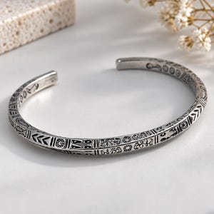 May include: A silver-toned metal cuff bracelet featuring an open-ended design. The exterior surface is decorated with a continuous pattern of engraved symbols, including small circles, geometric lines, and stylised animal figures in dark relief.