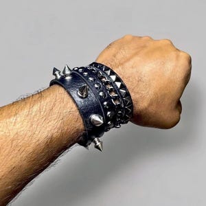 May include: A selection of black leather bracelets featuring silver spikes, studs, and chain elements. The bracelets are stacked on a wrist, displaying a punk rock or alternative aesthetic. The image is set against a neutral backdrop.