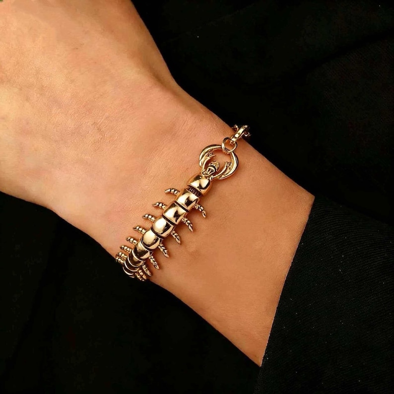 May include: A gold-toned bracelet featuring a segmented, articulated design that mimics the appearance of a centipede. The piece consists of multiple curved, metallic links with small, protruding legs along the sides and a circular clasp.