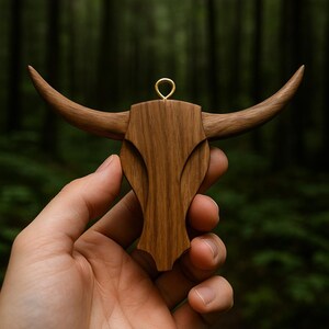 May include: A wooden bull skull pendant with a gold-colored ring for hanging. The pendant is carved from a light brown wood, featuring detailed horns and a textured surface. The background is a blurred forest scene.