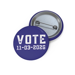 May include: A round pin-back button with a dark blue background. The front features the white text VOTE and the date 11-03-2026 in a distressed, block-style font. The back of the button shows a silver-toned metal pin mechanism for attachment.