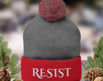 May include: A gray beanie with a red brim and a red pom-pom. The word "RESIST" is embroidered in white on the brim. The hat is displayed on a wooden surface with pine cones and greenery.