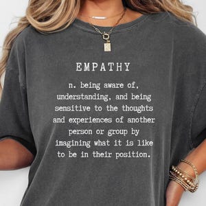 May include: Dark gray t-shirt with the word "EMPATHY" and its definition printed in white. The definition describes being aware of, understanding, and sensitive to the thoughts and experiences of others.
