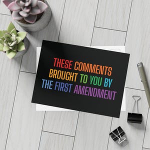 May include: Black postcard with rainbow text that reads "These comments brought to you by the First Amendment." The postcard is on a white surface with a pen, binder clips, and small potted plants.
