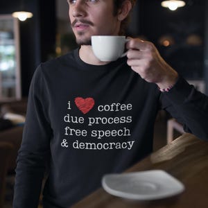 May include: Black long-sleeve shirt with the text "i ❤️ coffee due process free speech & democracy" in white. A red heart graphic is between the words "i" and "coffee". A white coffee cup is held up.