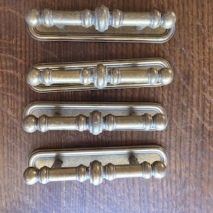 May include: Four antique brass drawer pulls. Each pull has a rectangular backplate with a decorative handle featuring a central design and rounded ends. The pulls are arranged on a wooden surface.