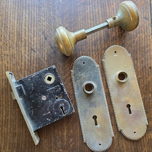 Puede incluir: Juego de herrajes de puerta de latón antiguo, que incluye un pomo, una cerradura y dos placas decorativas. El pomo tiene forma redonda con un borde detallado. La cerradura es negra y rectangular.