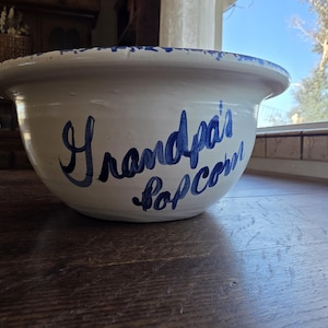以下が含まれることがあります： 青い縁と「Grandpa's Popcorn」の文字が青色で書かれた白い陶器のボウル。 ボウルは暗い木製の表面に置かれ、背景には窓と木が見えます。