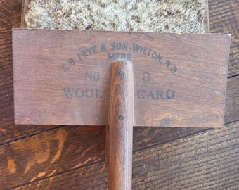 Könnte beinhalten: Eine Vintage-Wollkarte aus Holz mit Griff. Die Karte hat eine rechteckige Holzbasis mit dem Aufdruck "E.B. FRYE & SON WILTON, N.H. MFRS. NO. 8 WOOL CARD". Die Oberseite hat eine bürstenartige Oberfläche.