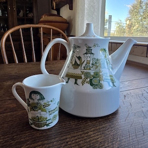 May include: White ceramic teapot and creamer set with a green and white illustration of figures carrying baskets. The teapot has a long spout and a curved handle. The creamer has a small handle and a similar design.