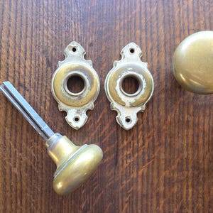 May include: Antique brass door hardware set, including two ornate backplates, a round doorknob, and a spindle with a matching knob. The hardware is set against a dark wood background.