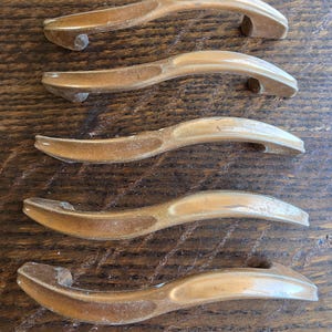 May include: Five vintage bronze-toned cabinet pulls. Each pull has a curved handle with a rounded end and a small, cylindrical post for mounting. The pulls are arranged on a dark wood surface.