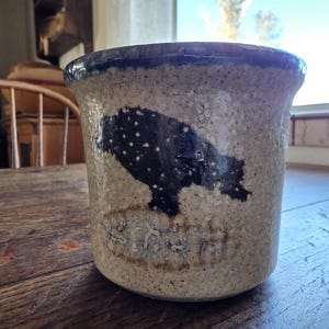 Puede incluir: Un tarro de gres moteado con un borde azul oscuro y una silueta de oso azul oscuro. El tarro tiene un color base marrón claro y una superficie texturizada. La palabra "YORK" está estampada en la base.