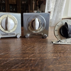Puede incluir: Tres temporizadores de cocina vintage. Dos son cuadrados, plateados, con esferas marcadas "Presto TIMER". El tercero es redondo, blanquecino, con una perilla negra y lee "MINUTE METER".
