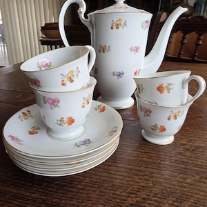 May include: A white porcelain tea set with floral designs. The set includes a teapot, teacups, and saucers. The cups and teapot have gold trim. The floral pattern includes orange, pink, and purple flowers. The set is arranged on a wooden table.